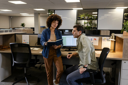 Diverse coworkers reviewing notebook at shared workstation in open-plan office, with dual monitors