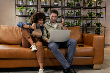 Browsing Diverse couple reviewing laptop on brown leather couch in living room, with coffee mugs