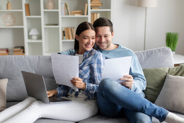 Happy young spouses paying bills online via laptop holding and reading papers, sitting on sofa,...