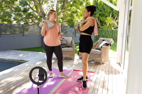 Chatting diverse female friends on wooden patio next to pool, with yoga mats and ring light
