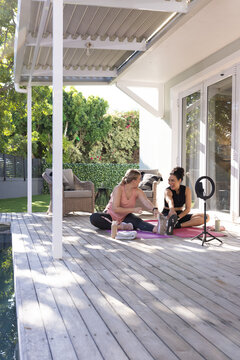 Stretching diverse female friends holding seated leg stretch on deck, with ring light and yoga mats