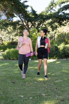Walking diverse female friends carrying yoga mats and water bottles on grassy lawn, with towels