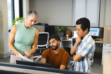 Diverse coworkers reviewing report at desk in modern office, with computer monitors and coffee cup