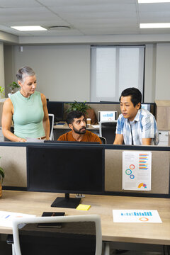 Discussing diverse coworkers reviewing charts at office desk, with wide monitor and sticky note