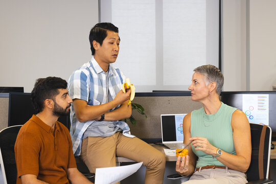 Taking break Diverse coworkers chatting in open-plan office, with banana fruit cup papers, monitors