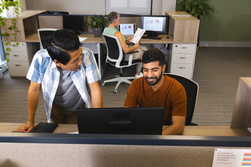 Collaborating diverse coworkers leaning over desk at office, with monitors and printed charts