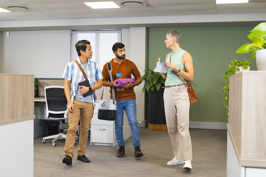 Walking diverse coworkers touring office corridor, with papers, yoga mat and water bottle
