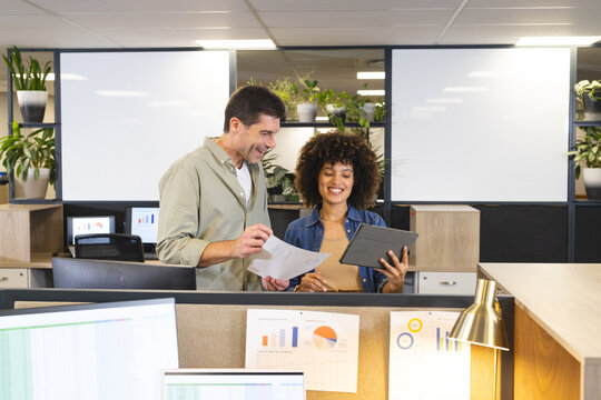 Diverse coworkers reviewing data in open-plan office, with tablet, printed charts and monitors