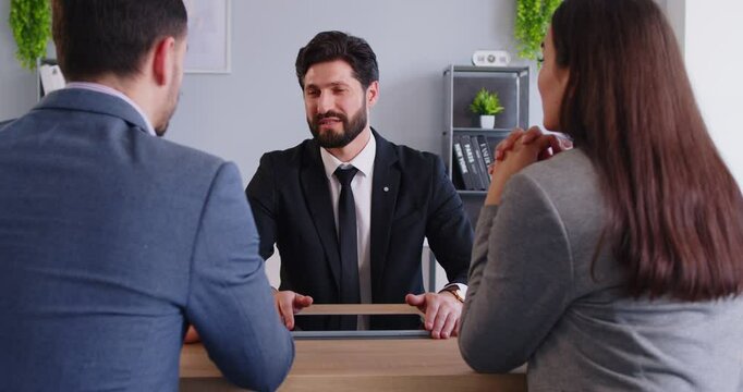 Male cheerful financial advisor consulting a young confident couple about insurance sitting at the desk in modern office. Man and woman talking with realtor or business broker. 4k video.