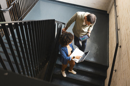 Working diverse coworkers analyzing data on laptop and tablet on office stairs with metal railings