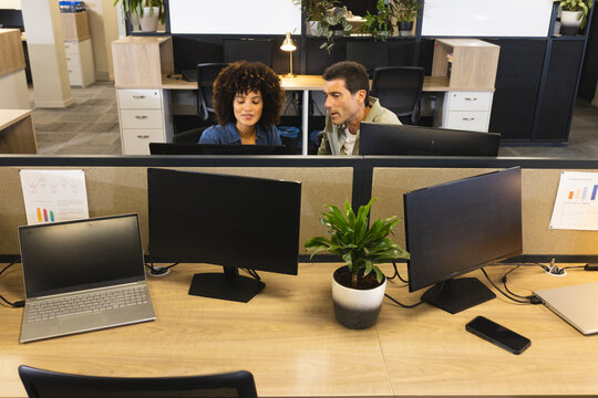 Collaborating Diverse coworkers reviewing data on two monitors at partitioned desks in office