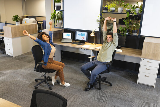 Leaning Diverse coworkers stretching arms overhead at office desk, with monitors and plants