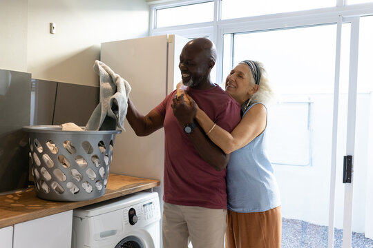 Laughing Diverse couple hugging and folding laundry in laundry room, with washing machine