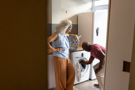 Senior couple bending down by front-loading washing machine in home laundry room, with woven basket