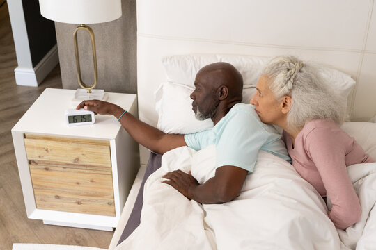 Diverse couple turning off digital alarm clock in bed, with bedside lamp and white bedding