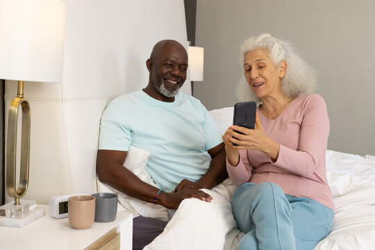 Showing diverse senior couple looking at smartphone on bed in bedroom, with lamp on bedside table