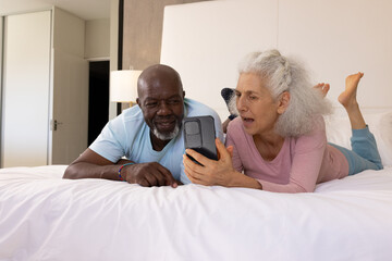 Laughing Diverse senior couple lying on bed with white linens in bedroom, with smartphone
