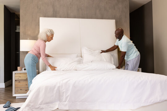 Smoothing duvet senior couple arranging king-size bed in master bedroom, highlighting plush pillows - Powered by Adobe