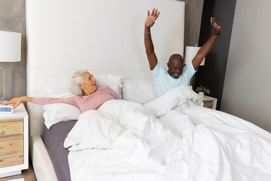 Stretching diverse senior couple rising under white duvet in bed, reaching for alarm clock