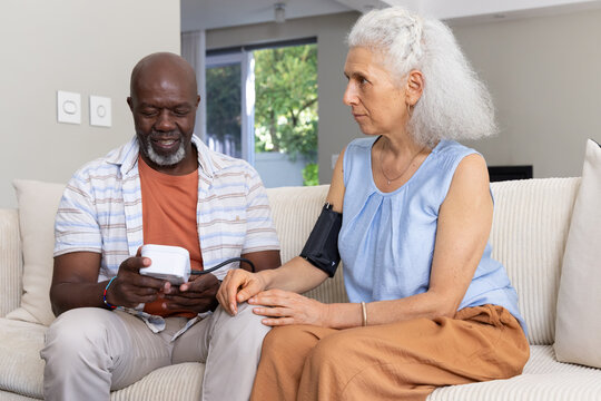 Measuring Diverse senior couple sitting on sofa in living room, using digital monitor and cuff
