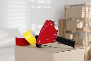 Dispenser with adhesive tape rolls on cardboard box indoors, closeup