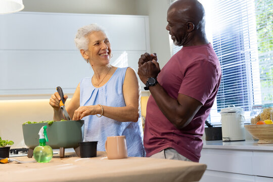 Senior couple stirring salad at kitchen counter, with mixing bowl spatula herb plant spray bottle