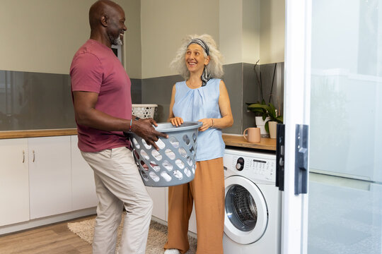 Diverse couple carrying laundry basket in home laundry room, with potted plants on countertop