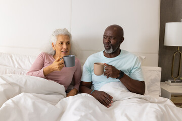 Sipping warm drinks senior couple sitting up in bed in bedroom, with ceramic mugs