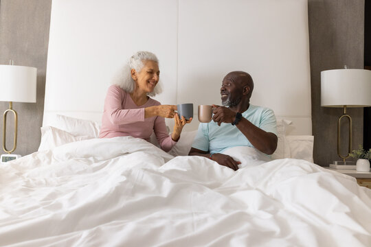 Clinking coffee mugs Diverse senior couple sitting in king-size bed at home with potted plant