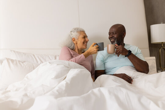 Clinking ceramic mugs Diverse senior couple sitting in bed at home, with white linens