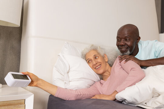 Reaching diverse senior couple lying in bed at home, with digital alarm clock on nightstand - Powered by Adobe