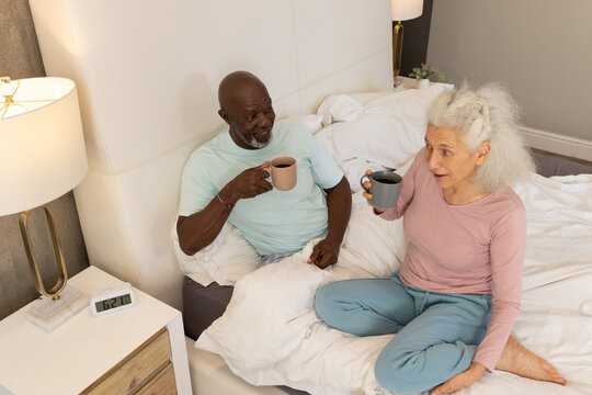 Sipping coffee Diverse senior couple sitting together on bed in modern bedroom, with ceramic mugs