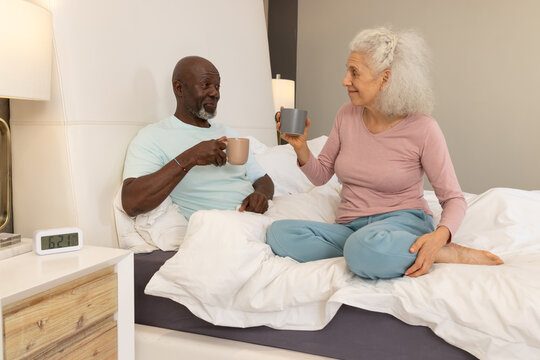 Holding mugs, diverse senior couple sitting on bed in modern bedroom, with digital alarm clock - Powered by Adobe