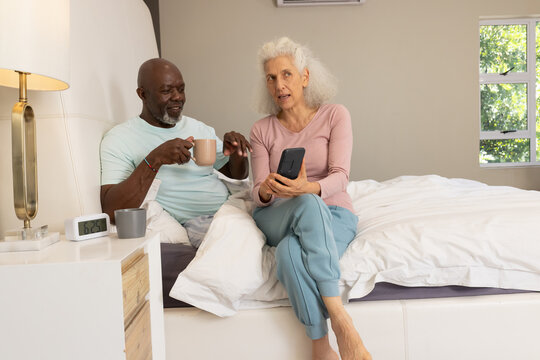 Relaxing diverse senior couple checking smartphone on bed, with coffee mugs, pillows and lamp