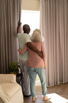 Embracing diverse senior couple standing in living room, with beige curtains and potted plant