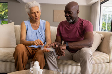 Diverse senior couple sorting pills into weekly pill organizer on sofa, with prescription bottles