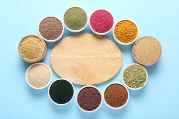 Different superfood powders in bowls and wooden board on light blue background, flat lay. Space for text