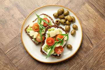 Delicious sandwiches with avocado, cheese, tomatoes, olives and arugula on wooden table, top view