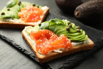 Delicious sandwiches with avocado, salmon, cream cheese, basil and capers on black textured table, closeup