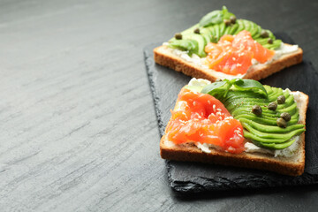 Delicious sandwiches with avocado, salmon, cream cheese, basil and capers on black textured table, closeup. Space for text