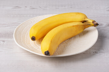 Yellow bananas on a white ceramic plate resting on a wooden table in a bright kitchen setting near a window
