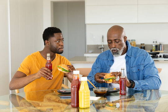 African American father and son eating burgers at glass-top table in kitchen, with soda and ketchup