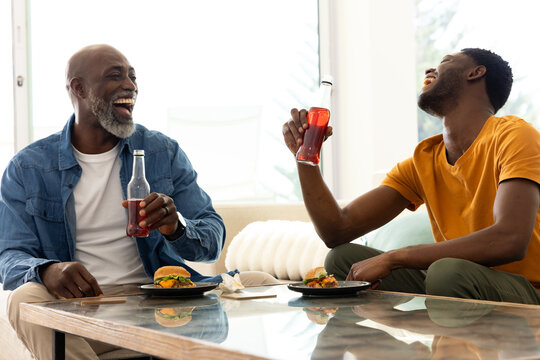 Laughing African American father and son raising red soda bottles in living room, with burgers