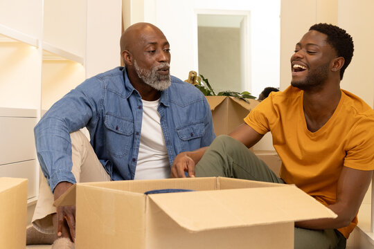 Laughing African American father and son unpacking moving boxes on floor in room, with potted plant