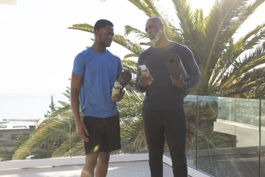 Standing African American father and son holding yoga mats and bottles on balcony overlooking ocean