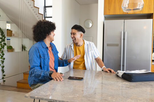 Laughing diverse male friends gesturing behind marble island in modern kitchen, with smartphone
