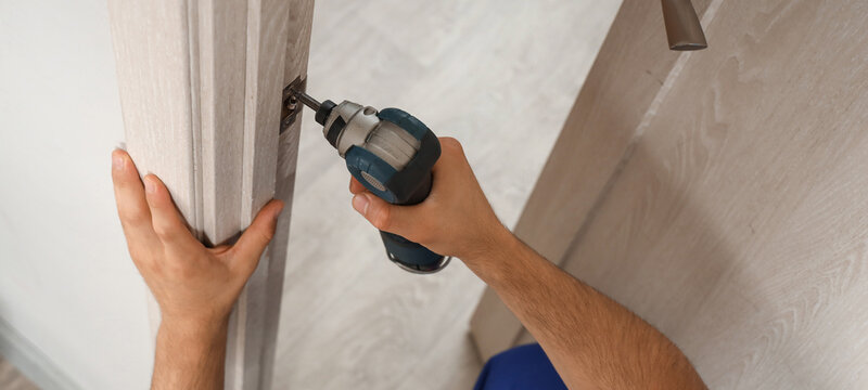 Male worker with electric drill installing door strike plate at home, closeup