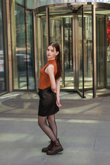 A girl with freckles in a leather skirt near a skyscraper.