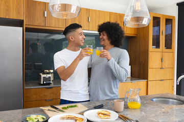 Diverse male friends toasting with orange juice in modern kitchen, with avocado and toast
