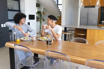 Diverse male friends grinding pepper onto avocado toast at kitchen dining table, with coffee mug